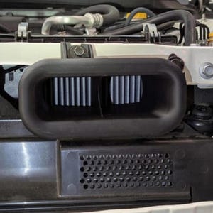 May include: A black automotive air intake system with two rectangular filter sections. The filters have vertical blue and white pleats. The system is installed in a vehicle's engine bay, with various hoses and components visible.
