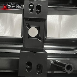 May include: Close-up of a black metal bracket with multiple holes and a circular silver accent. The bracket is part of a larger structure, with the text "V.2.STUDIO" visible in the upper left corner.