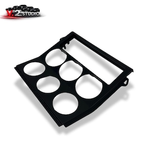 Peut inclure: Panneau de tableau de bord de voiture noir avec six ouvertures circulaires et un cadre rectangulaire. Le produit a une finition brillante et est conçu pour une utilisation automobile. Le logo "V.2 STUDIO" est visible.