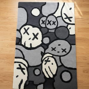 May include: A rectangular rug featuring a repeating pattern of cartoon faces in grey, black, and white. The faces have X-shaped eyes and mouths. The rug is placed on a wooden floor.