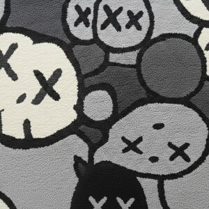 May include: Close-up of a rug with a repeating pattern of cartoon-like faces and shapes in shades of grey, black, and white. The design features stylized eyes and mouths, creating a playful and modern aesthetic.