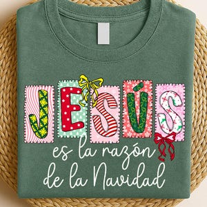 May include: Green t-shirt with the word "JESÚS" in colorful, decorative letters. The text "es la razón de la Navidad" is written below in white script. The shirt is displayed on a woven, circular mat.