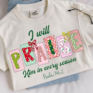 May include: Cream-colored t-shirt with the words "I will PRAISE Him in every season Psalm 146:2" in green and colorful block letters. The shirt is paired with blue jeans and gold-rimmed glasses.