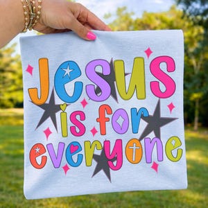 以下が含まれることがあります： 「Jesus is for everyone」とカラフルな泡文字で書かれた水色のTシャツ。星、ハート、十字架のデザインで、遊び心があり活気のある美しさです。