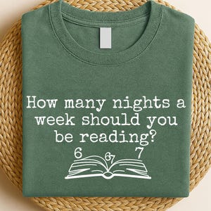 May include: Green t-shirt with the text "How many nights a week should you be reading?" in white. Below the text are the numbers 6, 6/7, and 7, along with an open book illustration. The shirt is displayed on a woven mat.