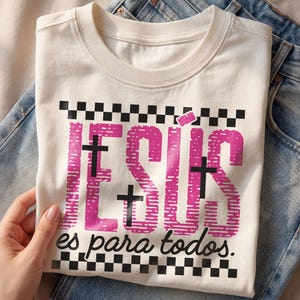 Cream Christian t-shirt folded over light wash denim jeans featuring bold pink distressed “JESÚS” lettering with integrated black crosses, checkerboard stripe accents, and the phrase “es para todos.” in script below. Styled in soft natural lighting with a hand holding the shirt, this faith-based Spanish graphic tee blends retro streetwear aesthetics with inspirational church apparel.