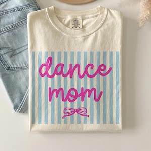 May include: Cream-colored t-shirt with the words "dance mom" in pink script over blue vertical stripes. A pink bow is below the text. Light blue jeans and a tan hat are also in the image.
