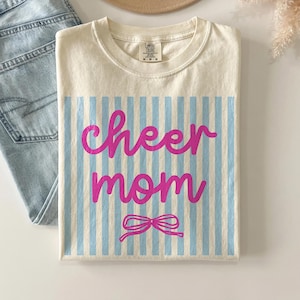 May include: Cream-colored t-shirt with the words "cheer mom" in pink script over a light blue striped background. A pink bow is at the bottom. The shirt is folded and next to a pair of blue jeans and a tan hat.