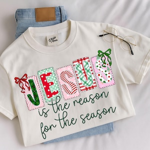 Könnte beinhalten: Ein cremefarbenes T-Shirt mit dem Wort JESUS in großen, gemusterten und bunten Blockbuchstaben, die mit roten und grünen Schleifen verziert sind. Darunter steht der Text is the reason for the season in dunkelgrüner Schreibschrift.