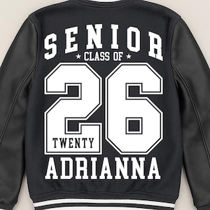Op de afbeelding: Zwarte varsity jas met witte letters en accenten. De achterkant toont "SENIOR", het jaar "26", de naam "ADRIANNA" en "CLASS OF". De jas heeft zwarte leren mouwen en witte strepen op de kraag, manchetten en zoom. Het jaar is voor het jaar 2026.