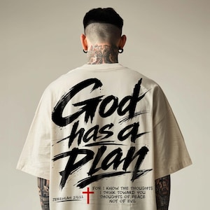 Back view of a cream oversized T-shirt worn by a male model against a clean neutral studio background. The shirt features a bold Christian streetwear graphic reading “God has a Plan” in large black brush lettering across the back, paired with a small red cross, Jeremiah 29:11 reference, and a shortened scripture message near the hem for a faith-based urban apparel look.