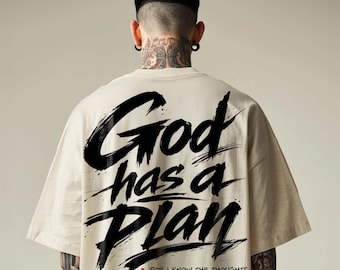 May include: A beige short-sleeved t-shirt featuring large black brushstroke text on the back that reads God has a Plan. Below the text is a small red cross and the scripture Jeremiah 29:11 with the text For I know the thoughts I think toward you thoughts of peace not of evil.