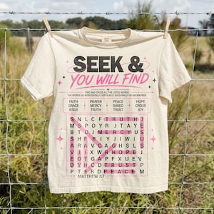 Cream Comfort Colors style T-shirt hanging on a wire fence outdoors, featuring a bold Christian “Seek & You Will Find” word search graphic in black and pink. The design includes faith words, Matthew 7:7 scripture, highlighted puzzle lines, and star accents, styled as a trendy faith-based shirt mockup with a rustic field background. Perfect for casual Christian apparel.