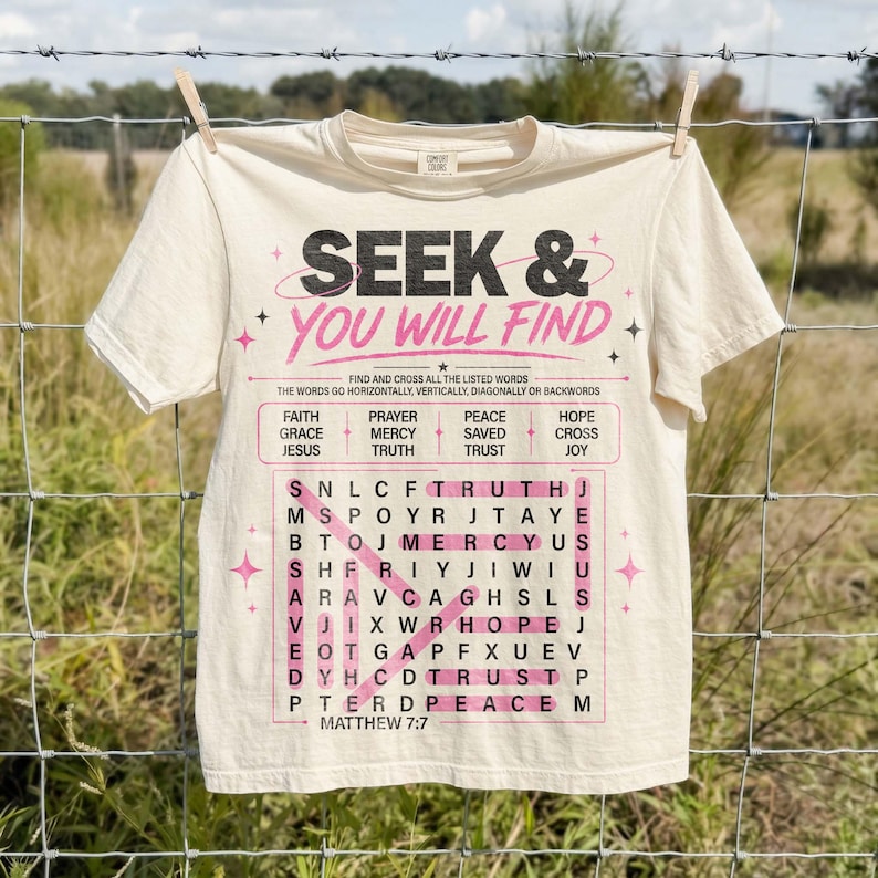 Cream Comfort Colors style T-shirt hanging on a wire fence outdoors, featuring a bold Christian “Seek & You Will Find” word search graphic in black and pink. The design includes faith words, Matthew 7:7 scripture, highlighted puzzle lines, and star accents, styled as a trendy faith-based shirt mockup with a rustic field background. Perfect for casual Christian apparel.