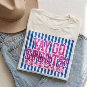 Styled flat lay of a neutral cream t-shirt placed on light denim with a beige hat and layered gold necklace, featuring a bold pink and blue “Yay Go Sports!” graphic with vertical stripes and motivational tagline “Do The Thing – Win The Points,” highlighting a sporty, preppy game day shirt mockup with modern feminine vibes.