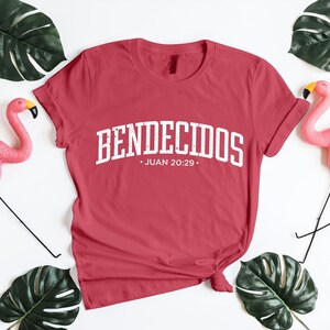 Styled flat lay mockup of a muted red unisex t-shirt featuring the Spanish word “BENDECIDOS” in large white distressed varsity lettering, with Juan 20:29 centered underneath. The shirt is neatly knotted and placed on a bright white background, framed by green monstera leaves and pink flamingo accents. This mockup emphasizes bold Christian typography, Spanish Bible verse apparel, and uplifting faith-inspired clothing visuals.