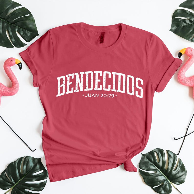 Styled flat lay mockup of a muted red unisex t-shirt featuring the Spanish word “BENDECIDOS” in large white distressed varsity lettering, with Juan 20:29 centered underneath. The shirt is neatly knotted and placed on a bright white background, framed by green monstera leaves and pink flamingo accents. This mockup emphasizes bold Christian typography, Spanish Bible verse apparel, and uplifting faith-inspired clothing visuals.