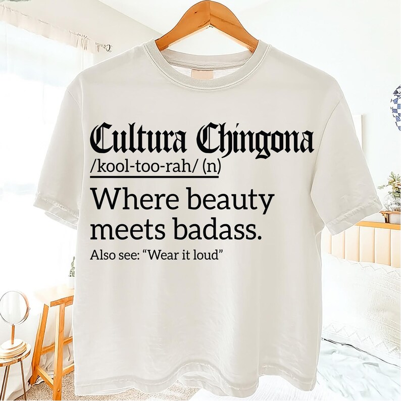 White T-shirt with bold black text reading "Cultura Chingona /kool-too-rah/ (n) Where beauty meets badass. Also see: 'Wear it loud'" in Gothic and serif fonts, displayed on a hanger indoors.