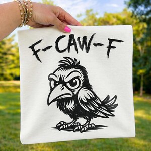 Hand holding folded cream t-shirt outdoors displaying bold black “F-CAW-F” brush typography and fierce crow artwork. Blurred green landscape background enhances this funny bird pun statement shirt mockup, highlighting the detailed feathers and expressive cartoon crow graphic.