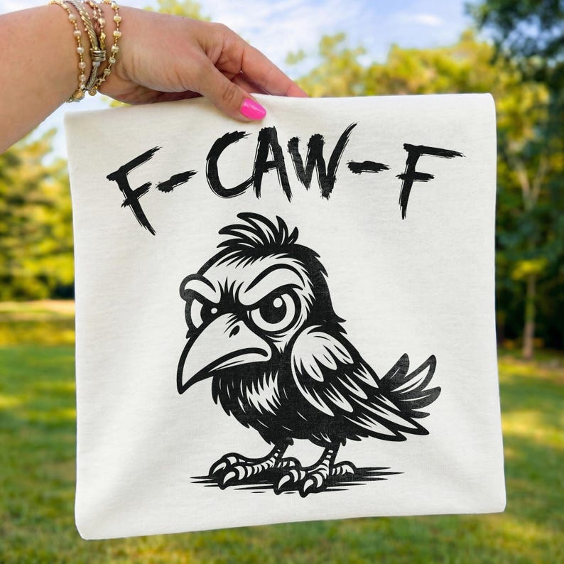 Hand holding folded cream t-shirt outdoors displaying bold black “F-CAW-F” brush typography and fierce crow artwork. Blurred green landscape background enhances this funny bird pun statement shirt mockup, highlighting the detailed feathers and expressive cartoon crow graphic.