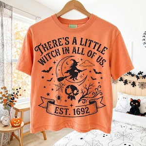 May include: An orange short-sleeved t-shirt featuring a black graphic print. The design includes a witch on a crescent moon, bats, a spider web, a skull, and bare branches. The text reads, There's a little witch in all of us. EST. 1692.
