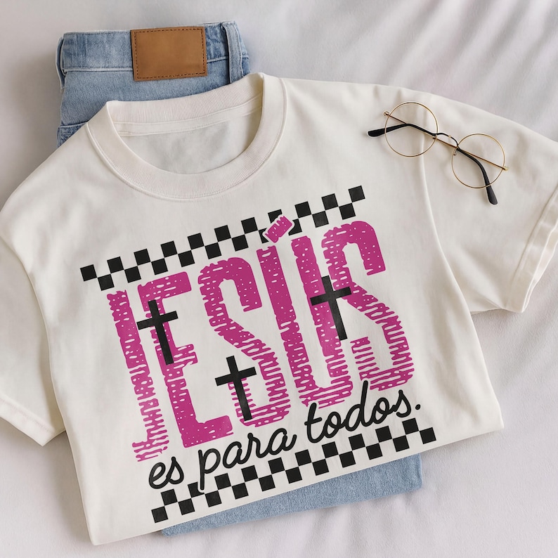 Cream Christian t-shirt folded over light wash denim jeans featuring bold pink distressed “JESÚS” lettering with integrated black crosses, checkerboard stripe accents, and the phrase “es para todos.” in script below. Styled in soft natural lighting with a hand holding the shirt, this faith-based Spanish graphic tee blends retro streetwear aesthetics with inspirational church apparel.