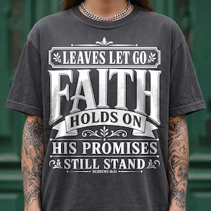 A model with tattooed arms wearing a dark washed charcoal gray t-shirt with a silver Cuban link necklace against a green background. The shirt features an intricate white vintage frame design reading "Leaves Let Go, Faith Holds On, His Promises Still Stand, Hebrews 10:23". A stylish fall-themed Christian graphic tee for casual everyday outfit looks.