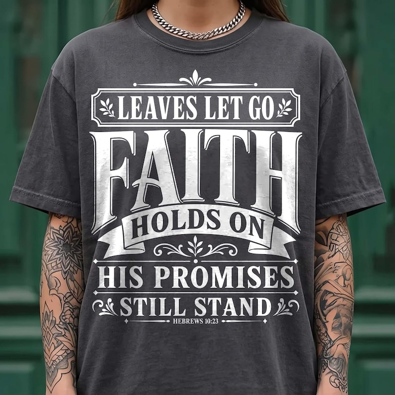 A model with tattooed arms wearing a dark washed charcoal gray t-shirt with a silver Cuban link necklace against a green background. The shirt features an intricate white vintage frame design reading "Leaves Let Go, Faith Holds On, His Promises Still Stand, Hebrews 10:23". A stylish fall-themed Christian graphic tee for casual everyday outfit looks.