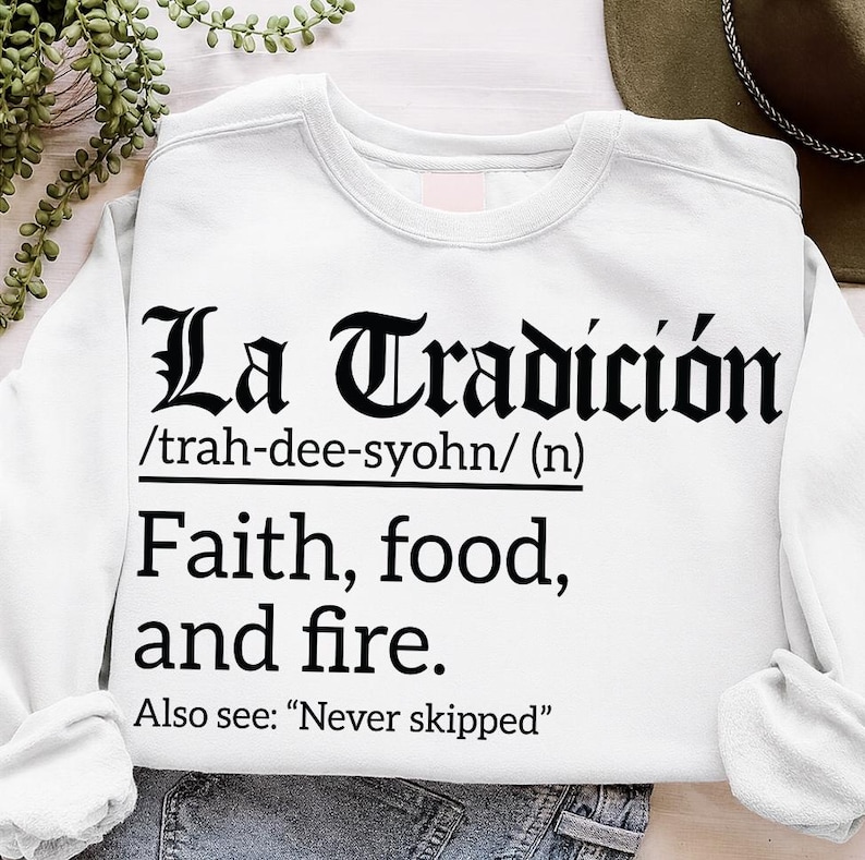White sweatshirt with bold black text reading "La Tradición /trah-dee-syohn/ (n) Faith, food, and fire. Also see: 'Never skipped'" in Gothic and serif fonts.