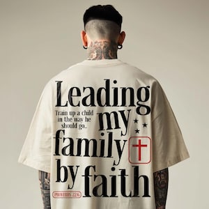 May include: Cream-colored t-shirt with the text "Leading my family by faith" in black, with a red cross graphic. The shirt also includes the Bible verse Proverbs 22:6.