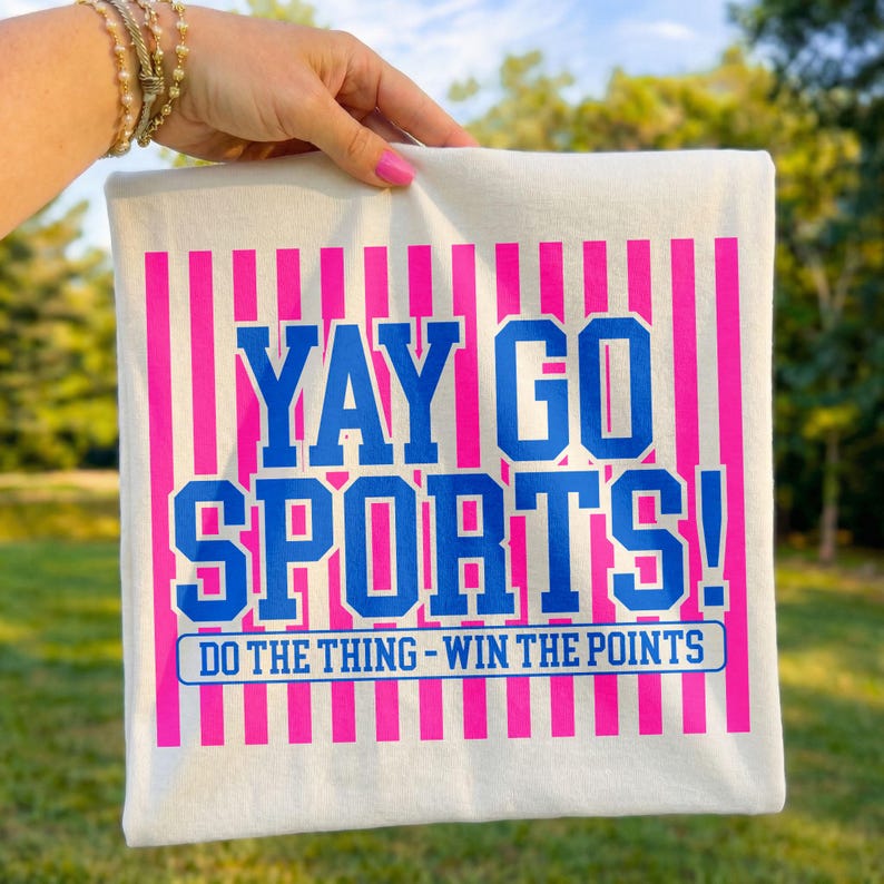 Outdoor lifestyle mockup of a folded white t-shirt held in hand with sunlit greenery background, featuring a bright pink striped backdrop and bold blue “Yay Go Sports!” varsity lettering with “Do The Thing – Win The Points” slogan, creating an energetic sports fan shirt presentation ideal for game day apparel displays.