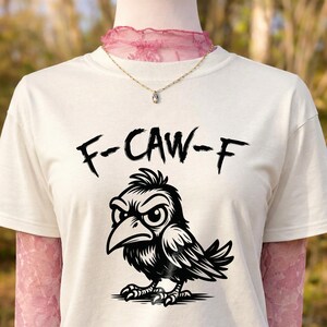 Close-up of cream t-shirt layered over pink lace long sleeves and styled with delicate necklace. Black brush-style “F-CAW-F” lettering sits above detailed cartoon crow artwork. Warm outdoor setting with blurred trees enhances this bold, sarcastic bird pun graphic tee presentation.