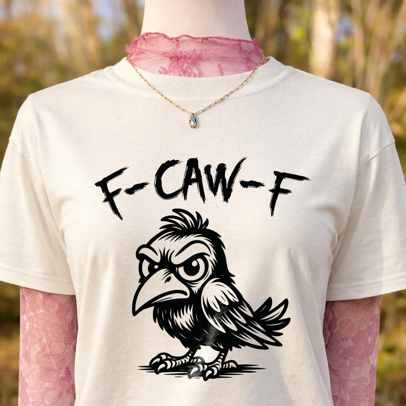 Close-up of cream t-shirt layered over pink lace long sleeves and styled with delicate necklace. Black brush-style “F-CAW-F” lettering sits above detailed cartoon crow artwork. Warm outdoor setting with blurred trees enhances this bold, sarcastic bird pun graphic tee presentation.