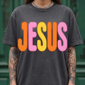 May include: A charcoal gray short-sleeve t-shirt featuring the word JESUS printed in large, scalloped-edge letters. The letters alternate between orange, pink, and yellow, each with a subtle vertical striped pattern throughout the design.