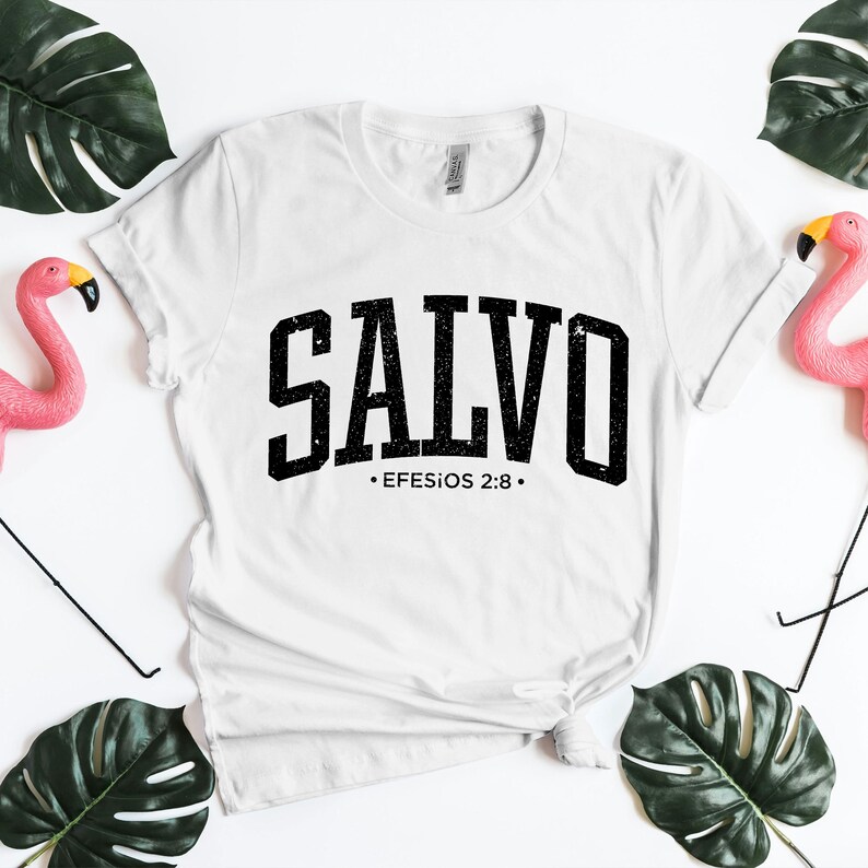 Clean flat lay of a white unisex Christian t-shirt showcasing the bold black distressed word “SALVO” in collegiate-style typography, with Efesios 2:8 printed below. The shirt features a knotted hem and is photographed on a bright white background with tropical green leaves and pink flamingo decor. This mockup is perfect for displaying Spanish faith designs, minimalist Bible verse shirts, and modern Christian apparel presentations.