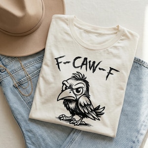 Flat lay of cream graphic t-shirt folded over light blue denim jeans with beige felt hat and gold necklace. Bold black brush lettering reads “F-CAW-F” above an angry cartoon crow illustration in detailed black ink style. Minimal neutral background and soft natural lighting create a trendy boutique mockup for this funny bird pun shirt design.