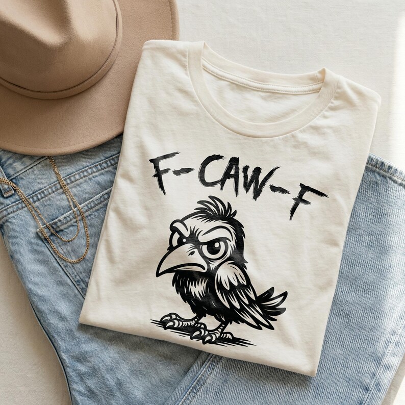 Flat lay of cream graphic t-shirt folded over light blue denim jeans with beige felt hat and gold necklace. Bold black brush lettering reads “F-CAW-F” above an angry cartoon crow illustration in detailed black ink style. Minimal neutral background and soft natural lighting create a trendy boutique mockup for this funny bird pun shirt design.