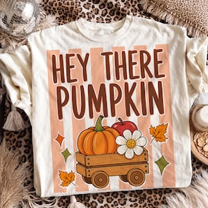Puede incluir: Una camiseta color crema con un gráfico que incluye el texto HEY THERE PUMPKIN en letras marrones. El diseño muestra un carro de madera con una calabaza naranja, una manzana roja y una flor blanca sobre rayas verticales color melocotón.