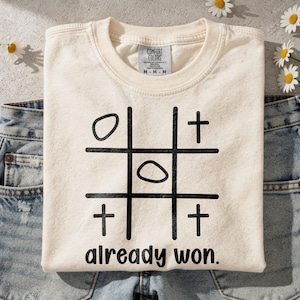 Könnte beinhalten: Cremefarbenes T-Shirt mit schwarzem Tic-Tac-Toe-Design und dem Schriftzug "already won." Das Shirt liegt gefaltet auf Jeans. Das Shirt hat ein Comfort Colors-Etikett.