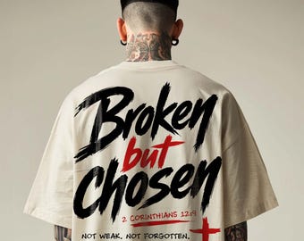 May include: A beige t-shirt featuring black and red graphic text on the back that reads Broken but CHOSEN, 2 Corinthians 12:9, NOT WEAK. NOT FORGOTTEN. HIS STRENGTH IS MADE PERFECT IN MY WEAKNESS, accompanied by a red cross symbol.