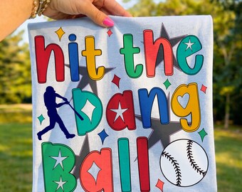 Op de afbeelding: Lichtblauw t-shirt met de tekst "Hit the Dang Ball" in kleurrijke letters. Het ontwerp bevat een honkbalspeler-silhouet, sterren en een honkbal. Een leuk en speels ontwerp voor honkbalfans.