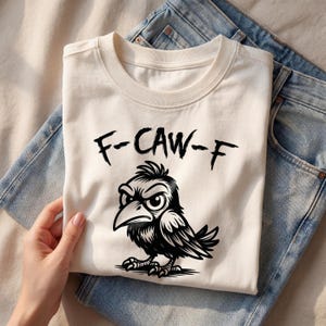 Top-down flat lay of cream graphic tee with bold black “F-CAW-F” lettering and detailed crow illustration, styled with denim jeans and neutral accessories. Clean beige backdrop and natural light emphasize the sharp linework and humorous bird pun design.