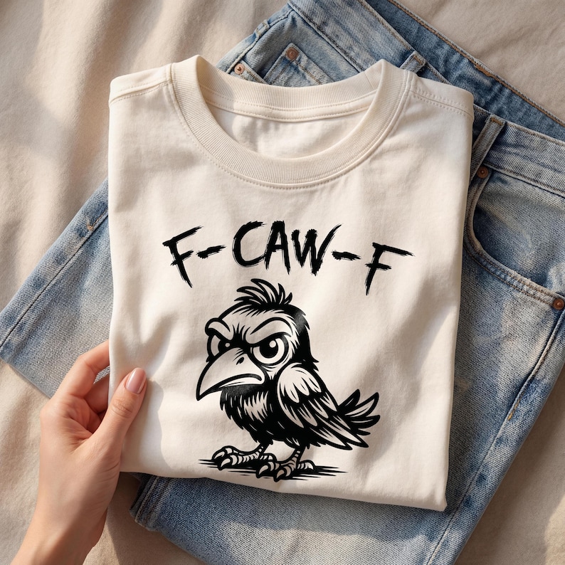 Top-down flat lay of cream graphic tee with bold black “F-CAW-F” lettering and detailed crow illustration, styled with denim jeans and neutral accessories. Clean beige backdrop and natural light emphasize the sharp linework and humorous bird pun design.