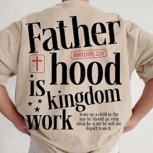 Back view of a man with short hair standing with his hands on his hips, wearing an oversized beige t-shirt and white pants. The back of the shirt shows a large Christian streetwear graphic with the bold text "Father hood is kingdom work" in black font. The design incorporates a red box with a Christian cross, a red Proverbs 22:6 capsule, three black stars, and the full text of the biblical child-rearing verse.