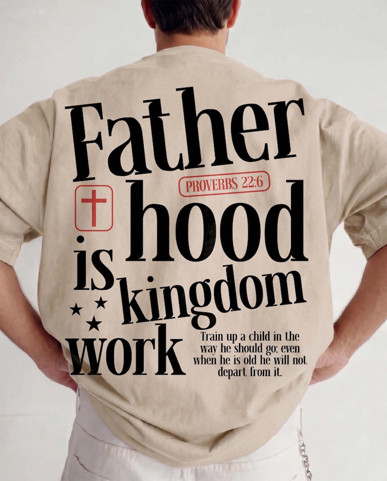 Back view of a man with short hair standing with his hands on his hips, wearing an oversized beige t-shirt and white pants. The back of the shirt shows a large Christian streetwear graphic with the bold text "Father hood is kingdom work" in black font. The design incorporates a red box with a Christian cross, a red Proverbs 22:6 capsule, three black stars, and the full text of the biblical child-rearing verse.