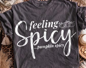May include: A charcoal gray t-shirt featuring white screen-printed text that reads "feeling Spicy" in a large script font, with the smaller text "...pumpkin spicy" below it and a small graphic of three pumpkins to the right of the word feeling.