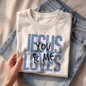 Close-up folded cream Comfort Colors t-shirt showing a bold vintage blue “Jesus Loves You & Me” Christian typography design with John 15:13 scripture printed on the front. A hand gently holds the shirt over light denim jeans, creating a warm lifestyle mockup scene. The distressed lettering emphasizes a trendy faith-based graphic tee suitable for Christian clothing listings and religious gift shirt presentations.