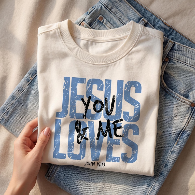 Close-up folded cream Comfort Colors t-shirt showing a bold vintage blue “Jesus Loves You & Me” Christian typography design with John 15:13 scripture printed on the front. A hand gently holds the shirt over light denim jeans, creating a warm lifestyle mockup scene. The distressed lettering emphasizes a trendy faith-based graphic tee suitable for Christian clothing listings and religious gift shirt presentations.