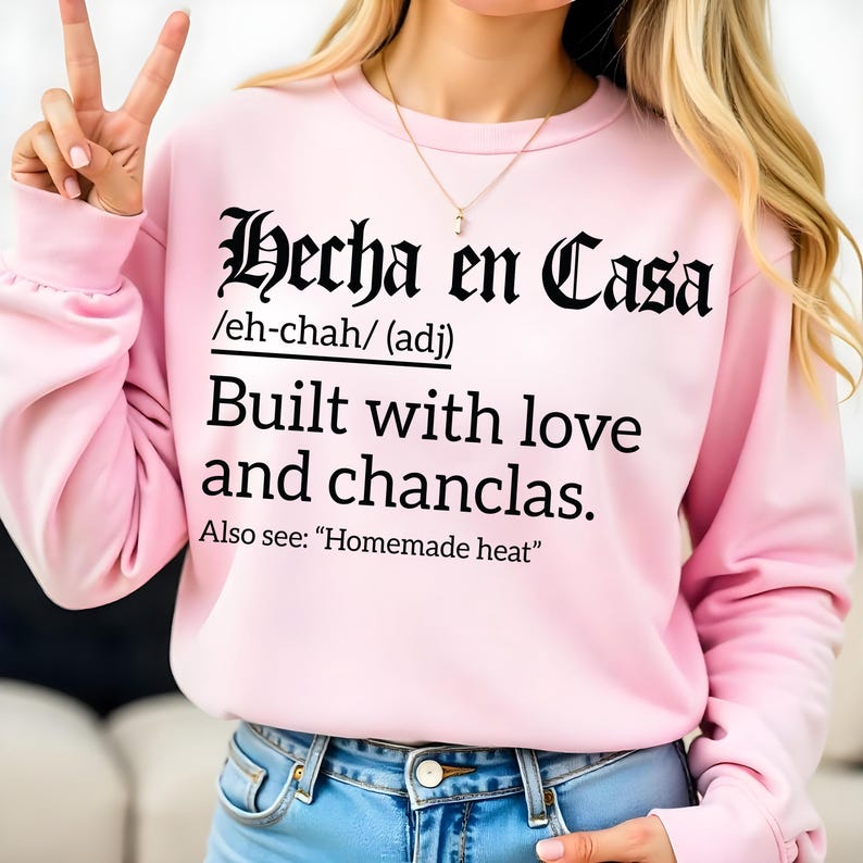 Light pink sweatshirt with bold black text reading "Hecha en Casa /eh-chah/ (adj) Built with love and chanclas. Also see: 'Homemade heat'" in Gothic and serif fonts, worn by a woman making a peace sign.