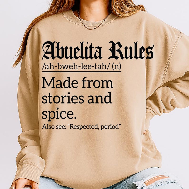 Beige sweatshirt with bold black text reading "Abuelita Rules /ah-bweh-lee-tah/ (n) Made from stories and spice. Also see: 'Respected, period'" in Gothic and serif fonts, worn with ripped light blue jeans.